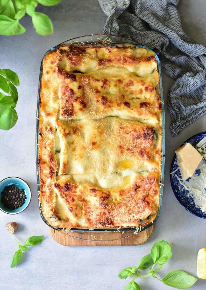 Vegetarian lasagna with zucchini and spinach in a baking dish. Parmesan cheese, kitchen cloth and basil leaves on the side.