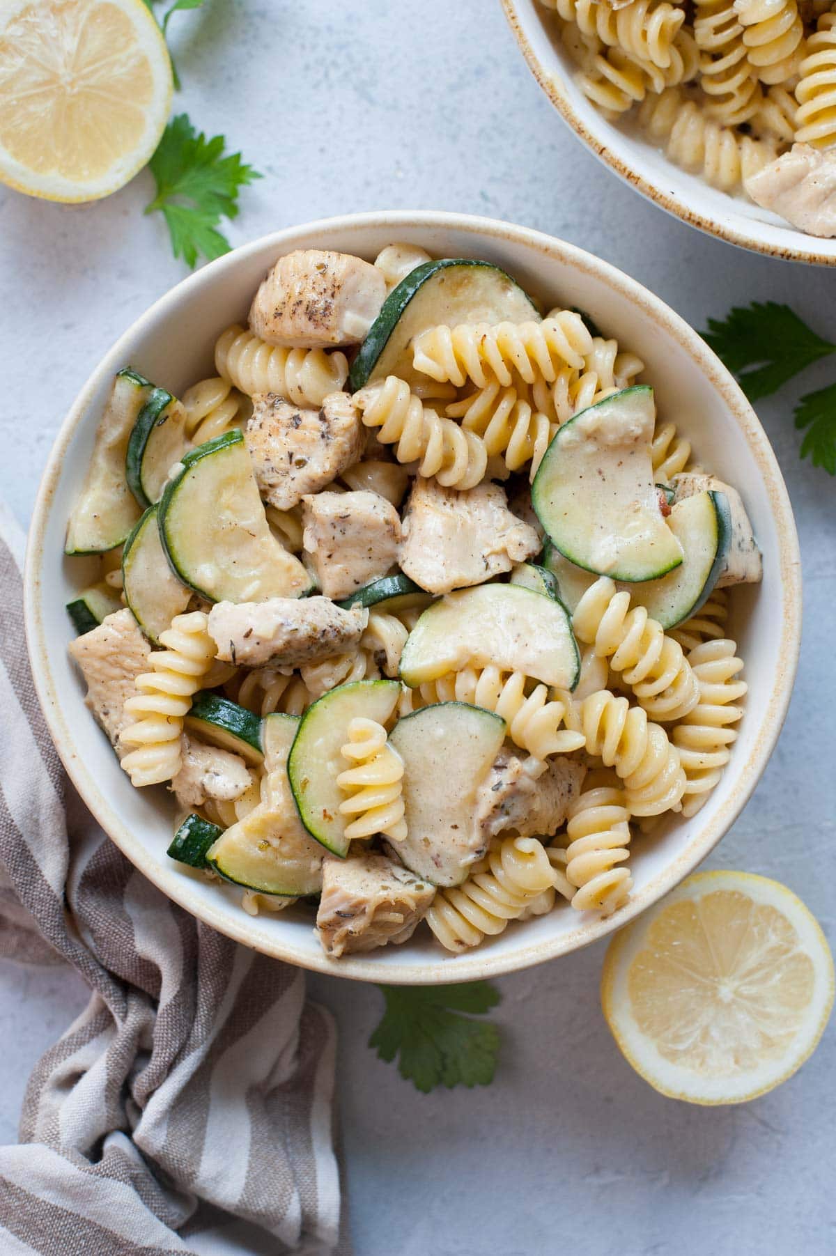 Chicken zucchini pasta in a white bowl. Lemons, parsley, and a kitchen cloth in the background.