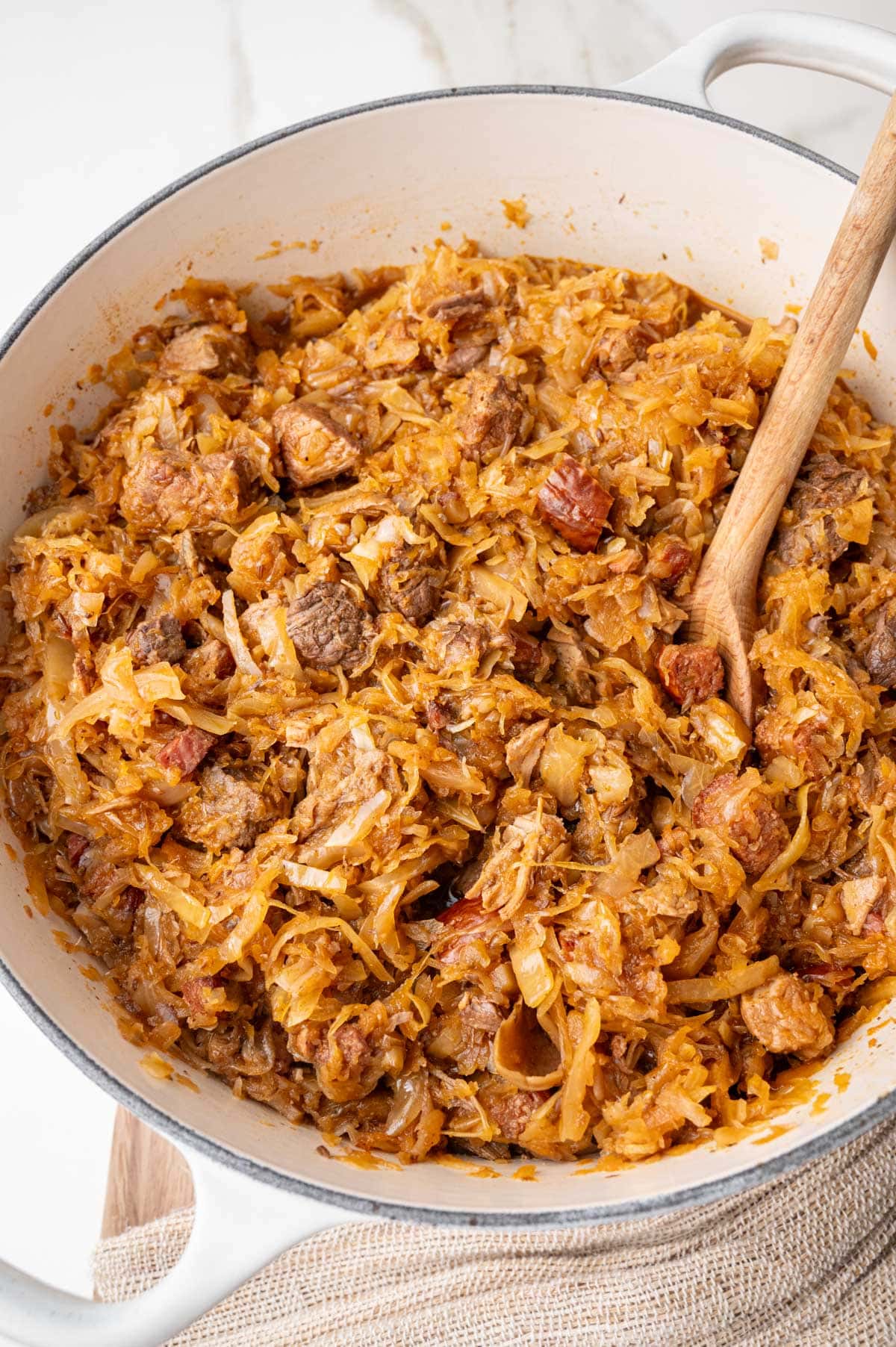 Bigos stew in a big white Dutch oven with a wooden spoon inside the pot.